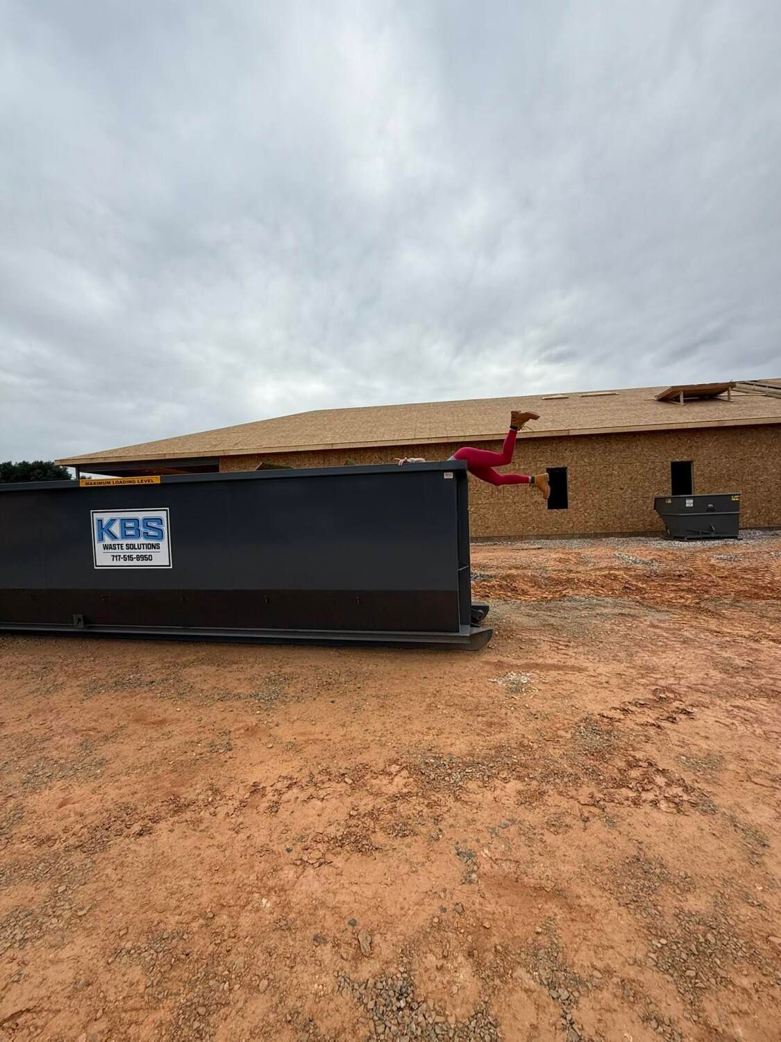 Person jumping over a black dumpster at a construction site. Red attire. Cloudy sky.