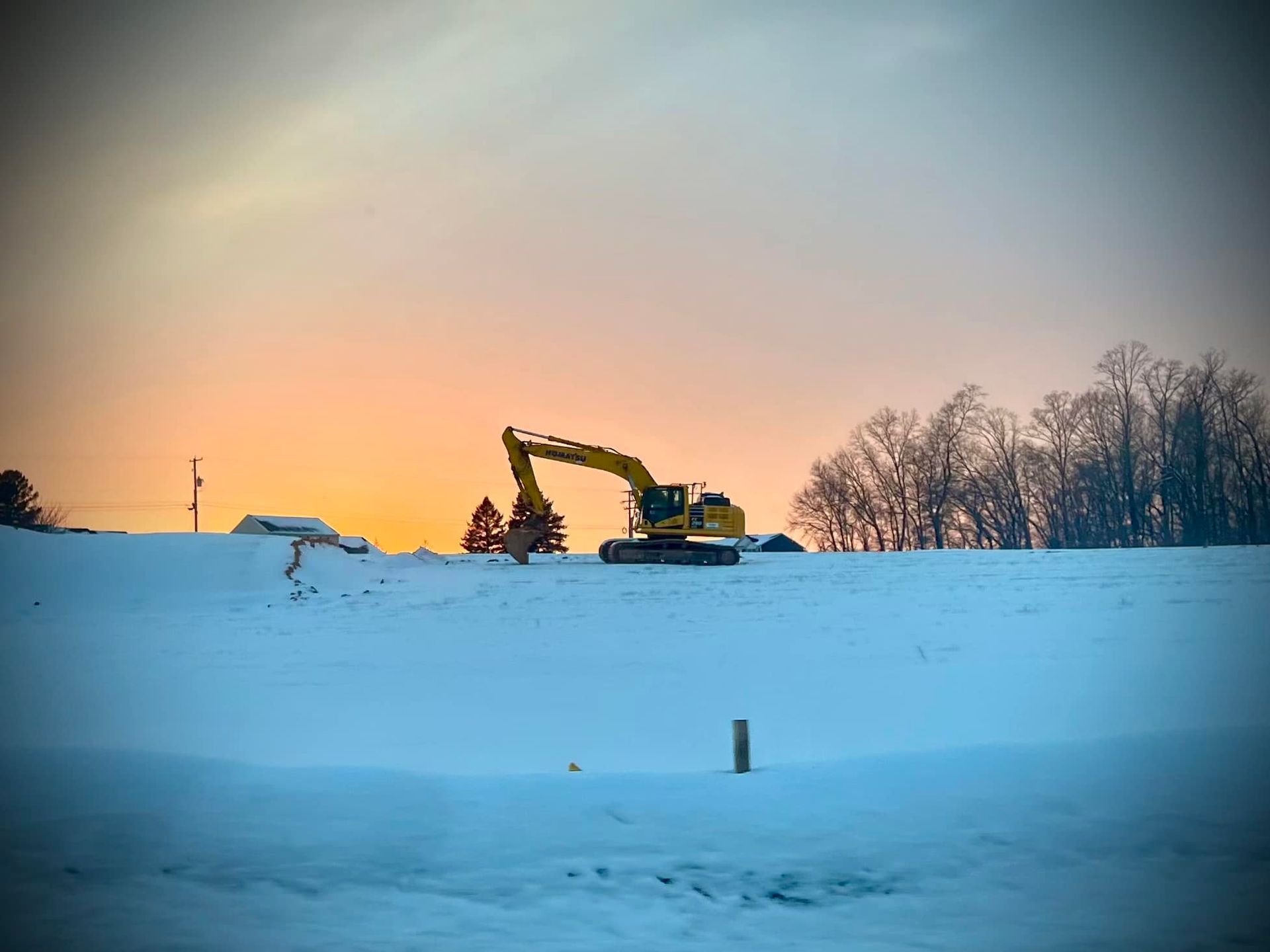 Excavator on snow-covered hillside silhouetted against a colorful sunset.
