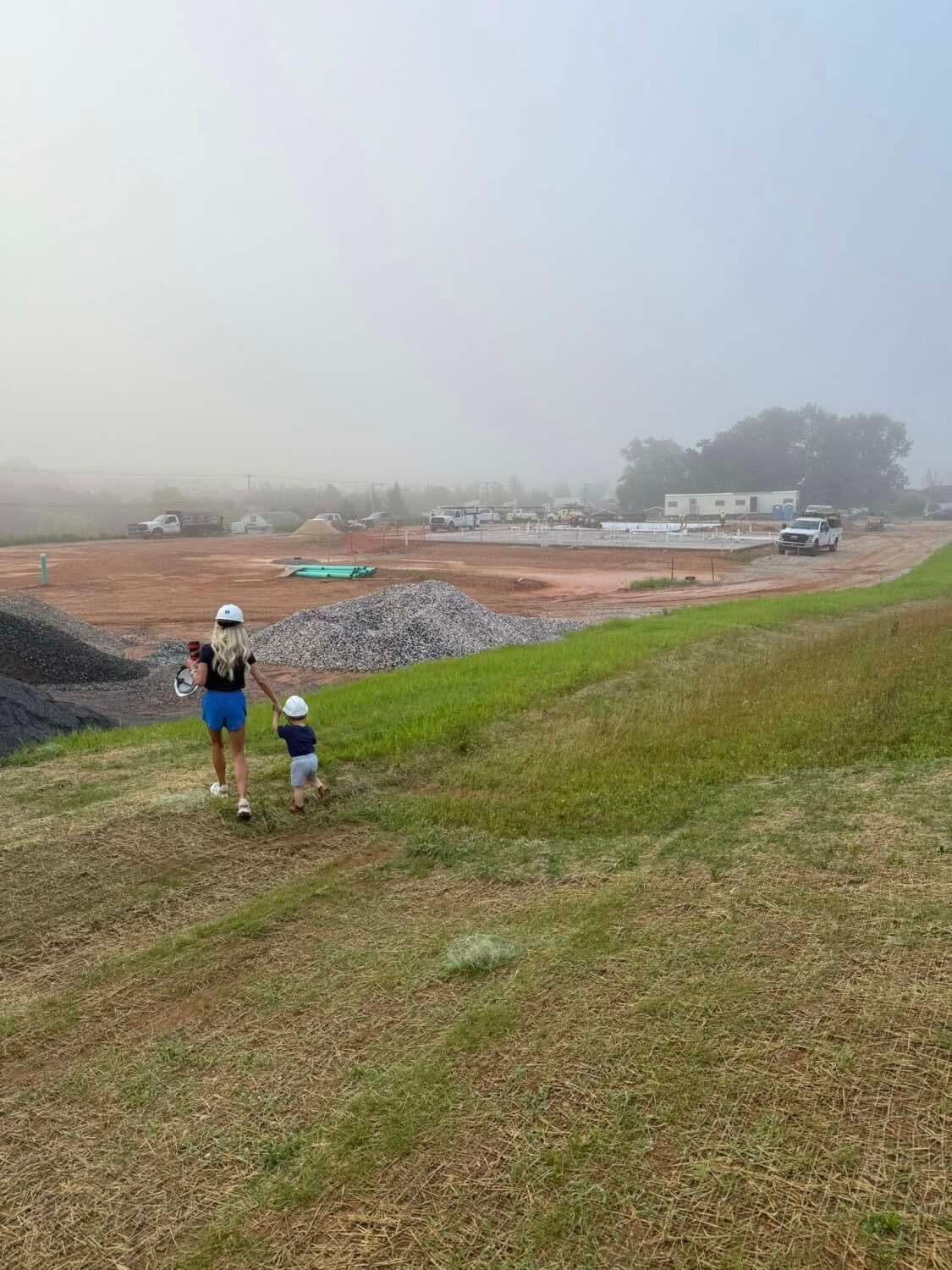Woman in blue shorts and child walk on grassy hill toward construction site in fog.