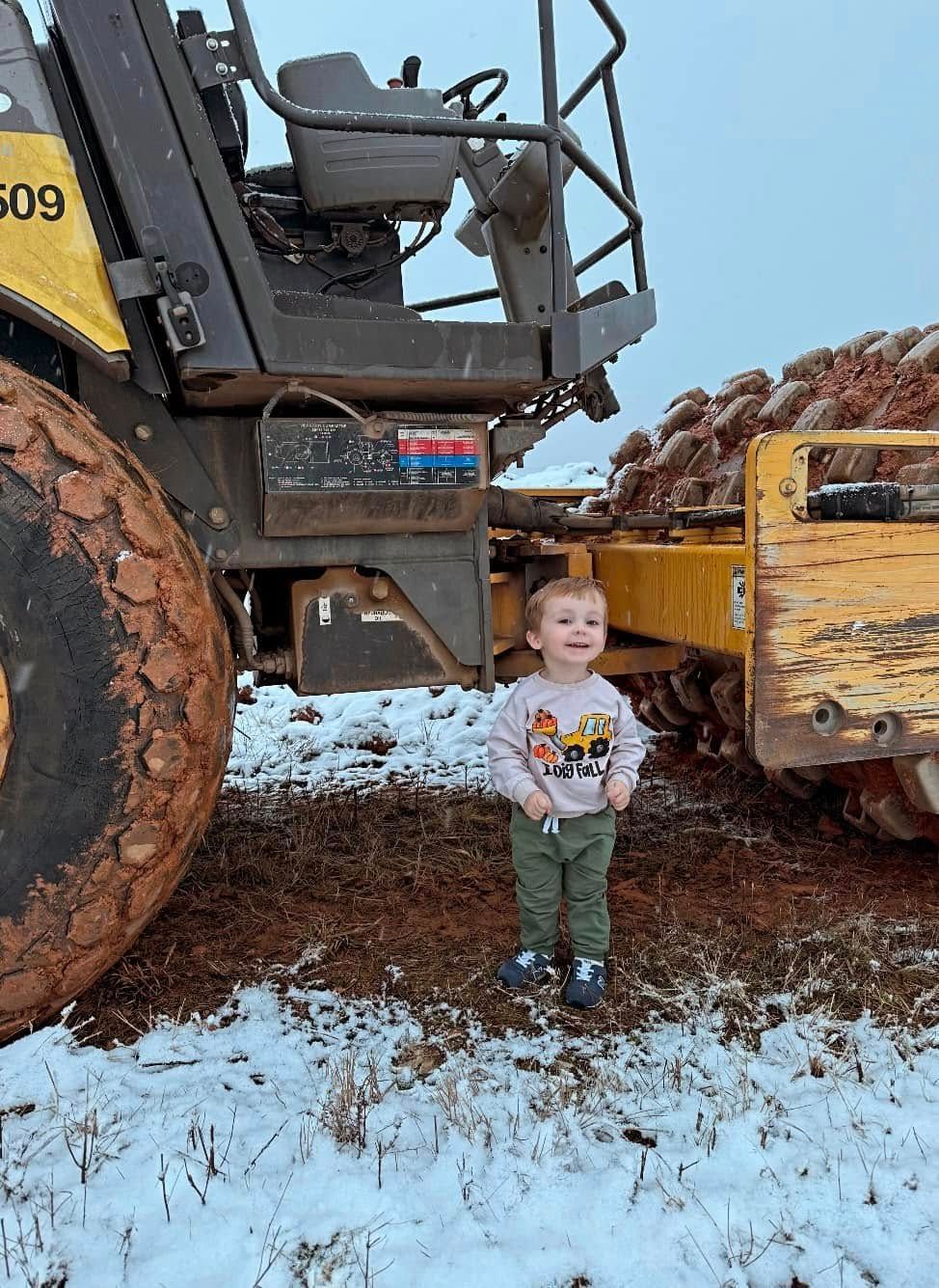 Young child stands smiling in front of construction vehicle, muddy ground with patches of snow.