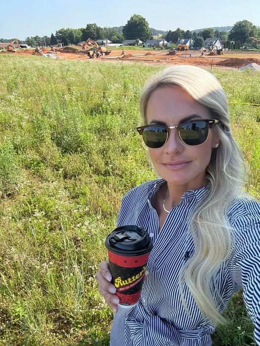 Woman holding coffee, standing in a field with construction in the background. She is wearing sunglasses and a striped shirt.
