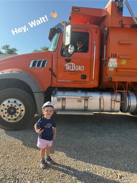 Young child in shorts waves at a construction truck driver in the cab. Orange truck reads