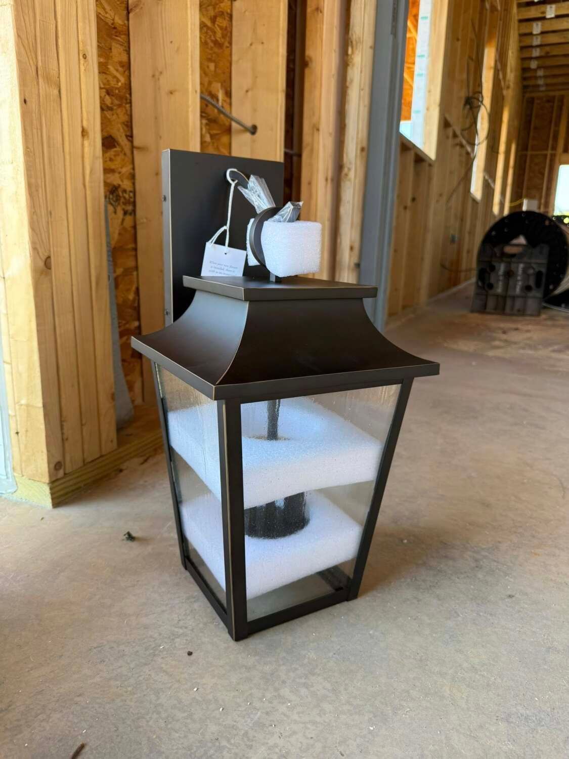 Black outdoor lantern, installed on a wooden wall, with light fixture visible inside, in a construction setting.