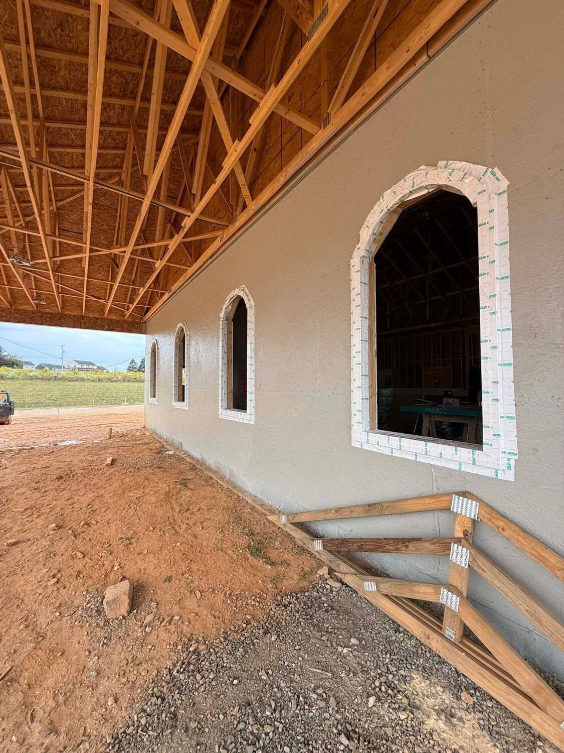 Exterior view of a building under construction. Stucco wall with arched windows; exposed wooden roof. Dirt ground.