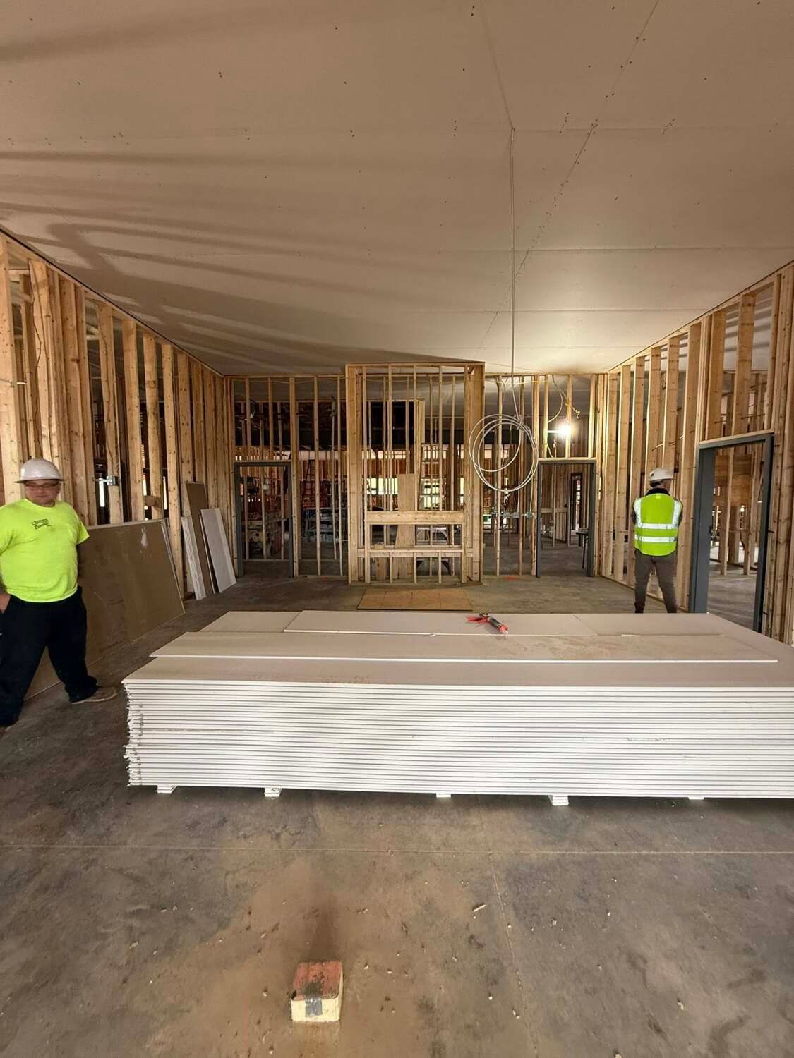 Construction site with exposed wooden framing and drywall. Two workers in safety vests.