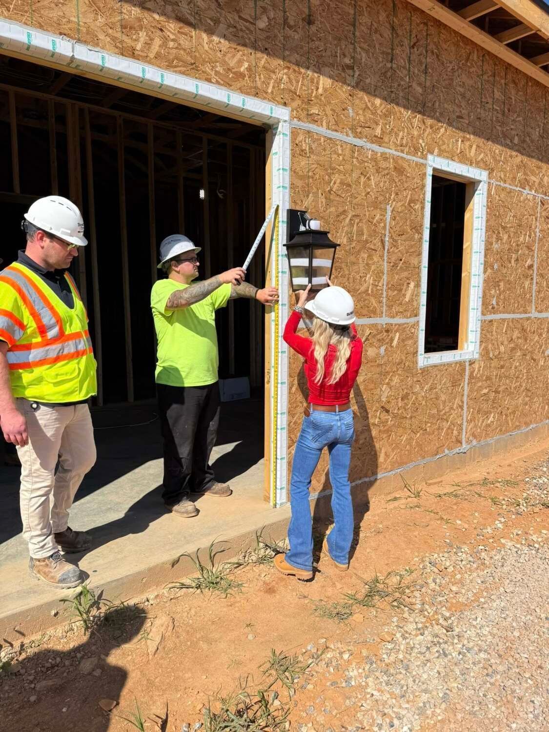 Three people at a construction site measure a doorway. One wears a hard hat and red top, another in a green shirt points with a ruler.