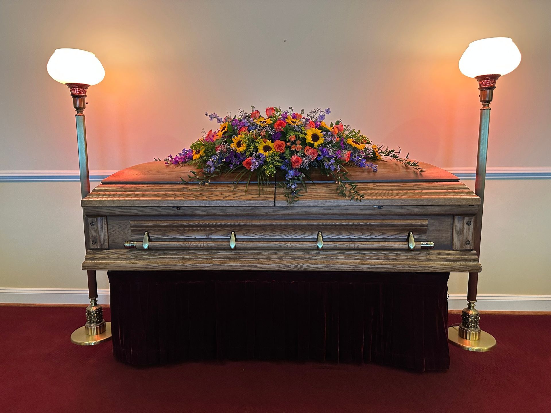 A wooden casket with floral arrangement, flanked by lamps, on a maroon-skirted stand, in a room with a red carpet.