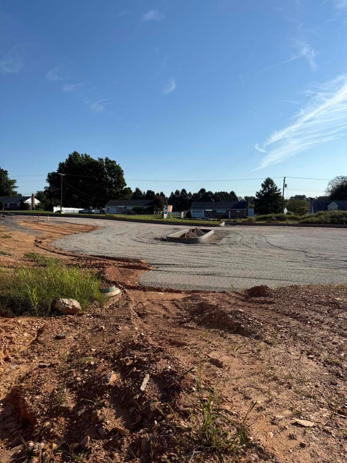 Dirt foreground with gravel and cracked pavement, low houses, trees, blue sky.