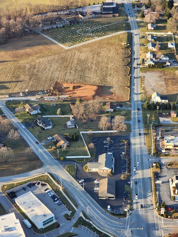 Aerial view of a road lined with houses and businesses, with a field and cemetery on the left.
