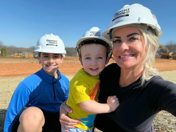 Woman and two children in construction hats smile on a sunny construction site.