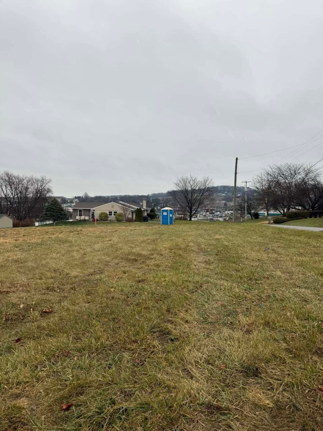 A field with a blue portable toilet in the center, houses and overcast sky in the background.