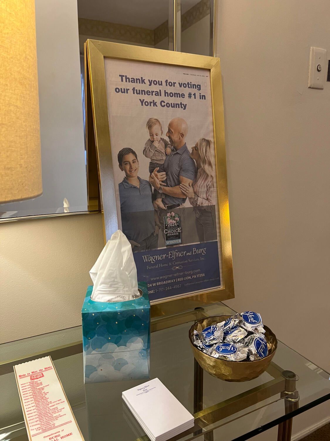 A framed sign with a family photo on a glass table. Next to it are tissues, candy, and a notepad.