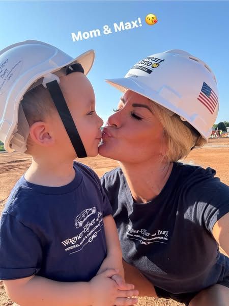 Mom and child in hard hats kiss. They are outside on a construction site.