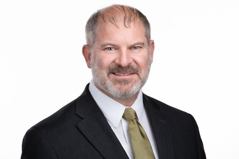 Man in suit smiles, looking at the camera. He has a beard and short hair, against a white backdrop.