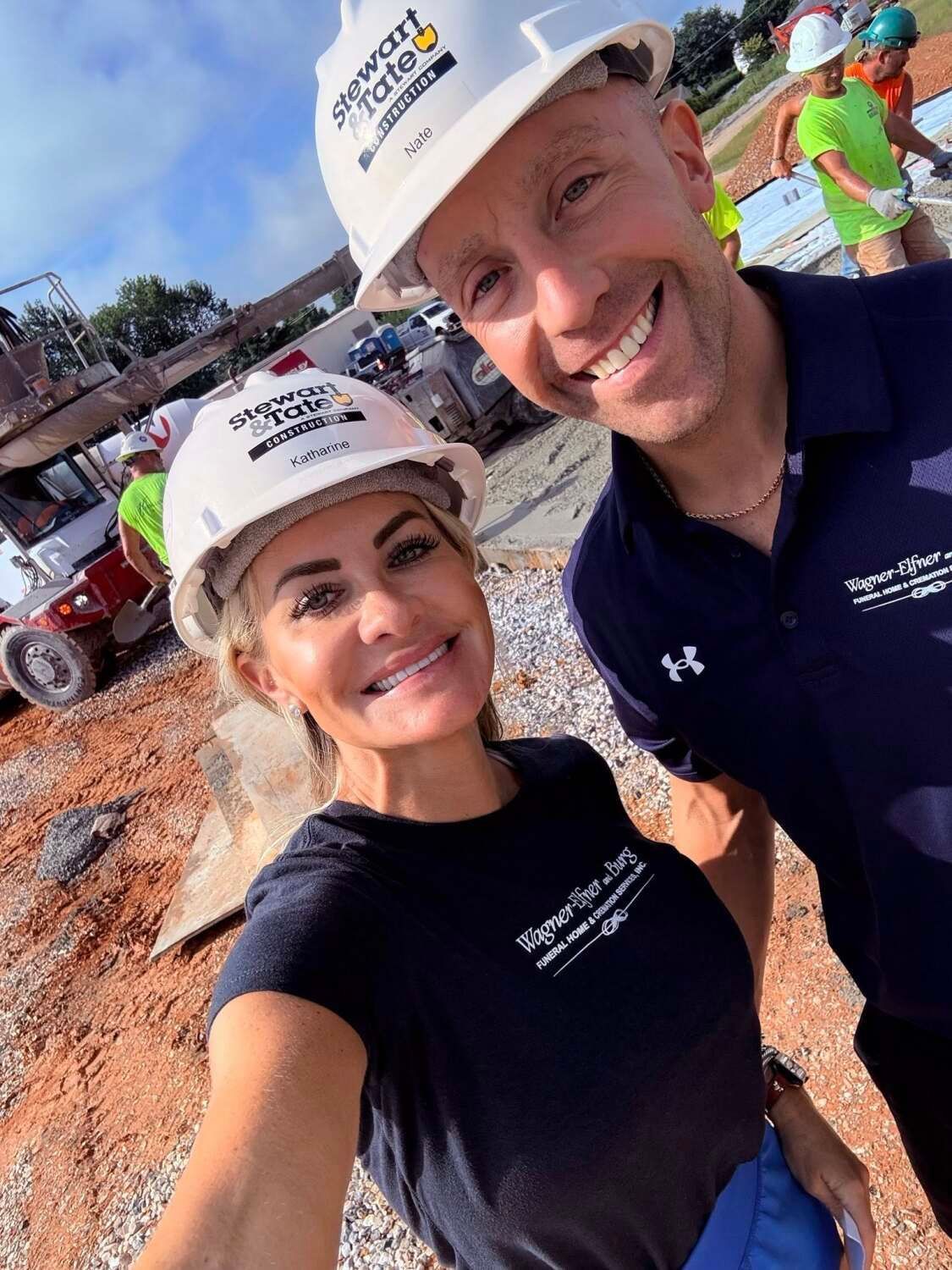 Two people in hard hats smile for a selfie at a construction site.