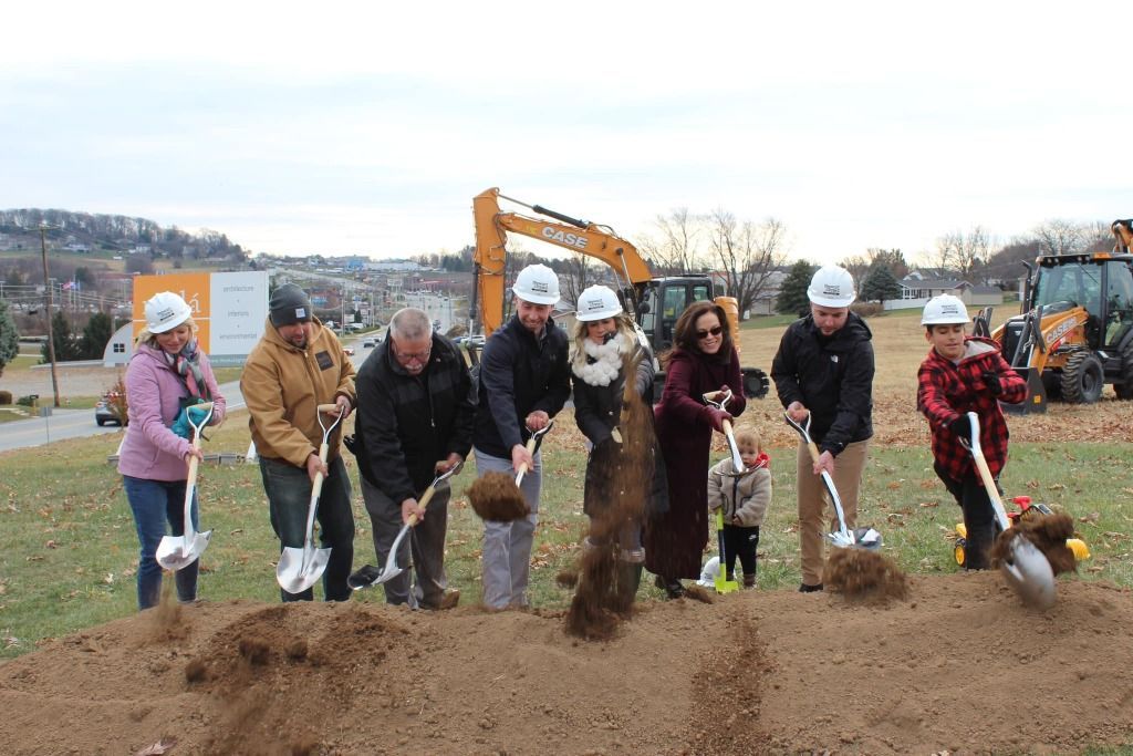 People with shovels at a groundbreaking ceremony, digging into a mound of dirt with construction equipment in background.