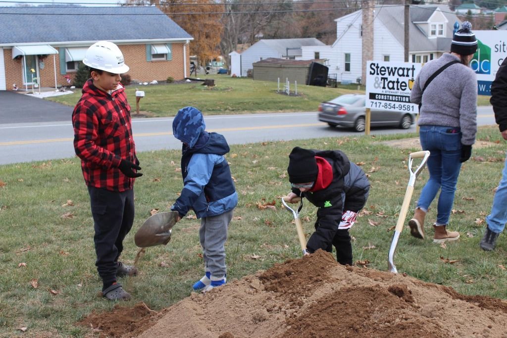 Children digging in a dirt pile with shovels at a ground-breaking event, wearing jackets and a hard hat.