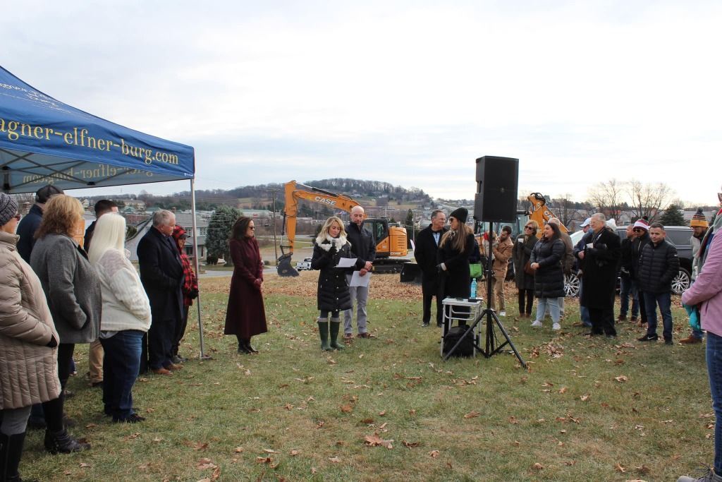 People gathered outdoors, listening to speakers near a tent. An excavator is visible in the background.