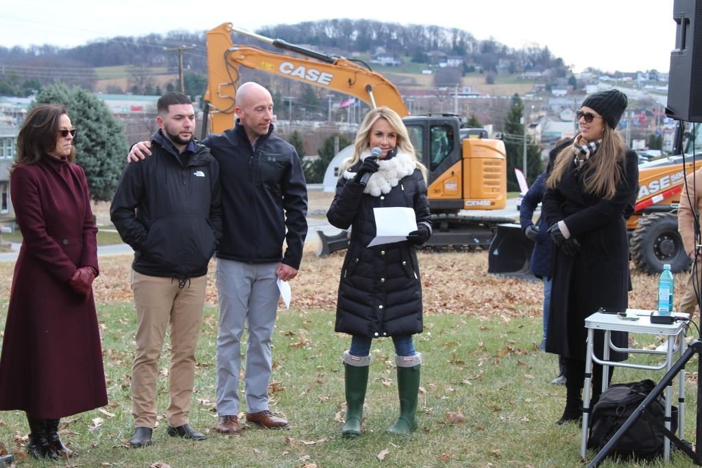 Group of people at an outdoor event with an excavator in the background, a woman speaking into a microphone.