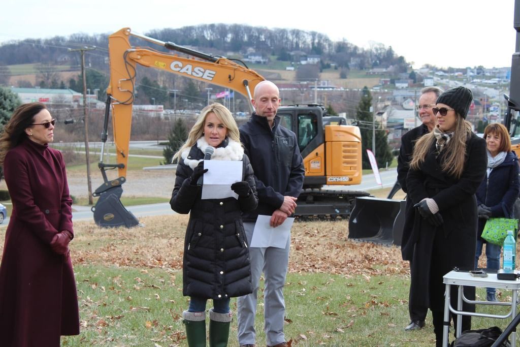People gathered outdoors, woman speaking, excavator in background, hill in distance.