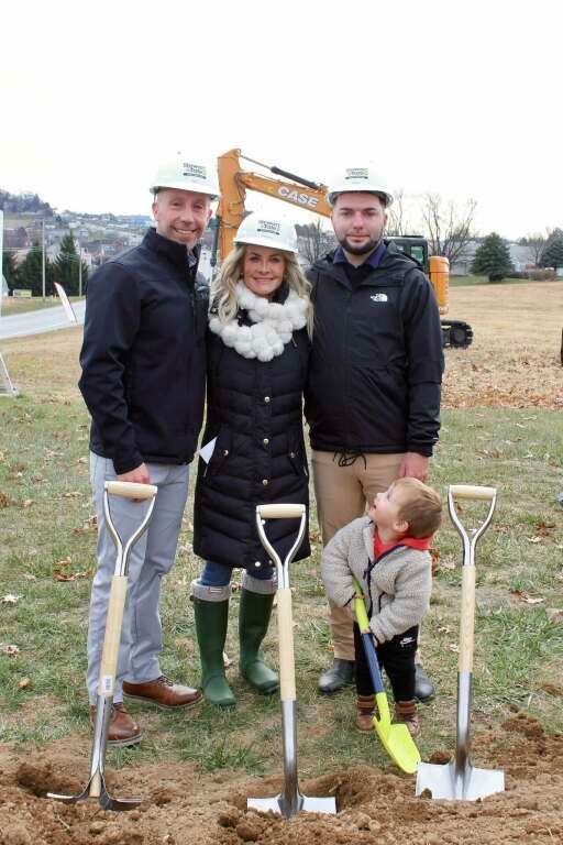 People at a groundbreaking ceremony with shovels; child digs with small shovel, excavator in background.