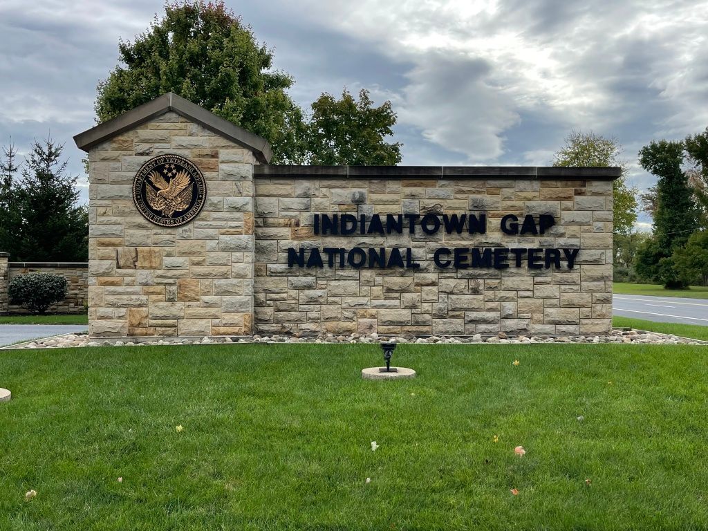 Sign for Indiantown Gap National Cemetery; stone wall, black text, cloudy sky.
