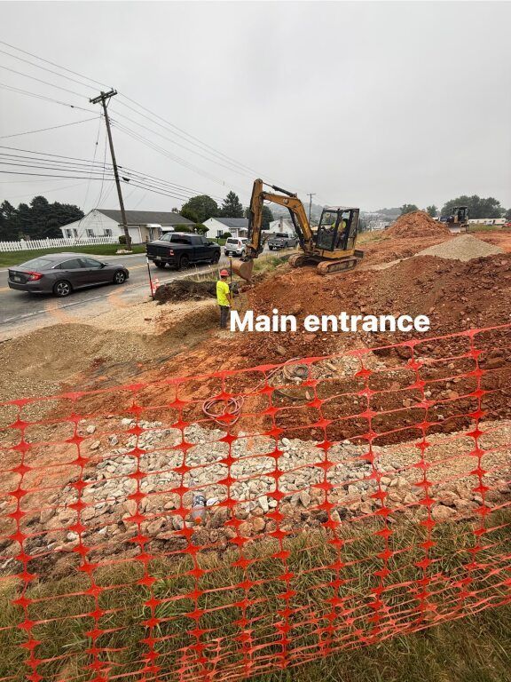 Construction site with excavator near main entrance, orange safety fencing, vehicles on road.