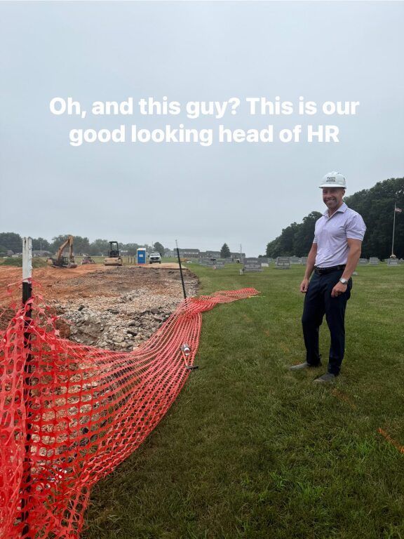 Man in hard hat stands by construction site, smiling. Red safety fencing and cloudy sky visible.