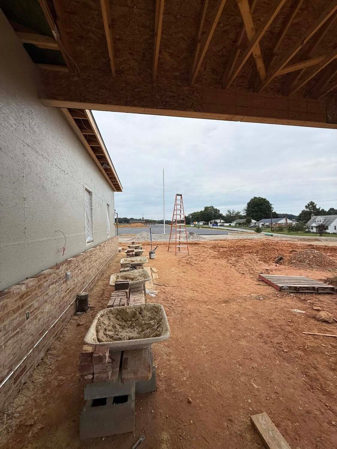 Construction site: exterior wall under a roof, wheelbarrow, dirt, and view of a residential street.