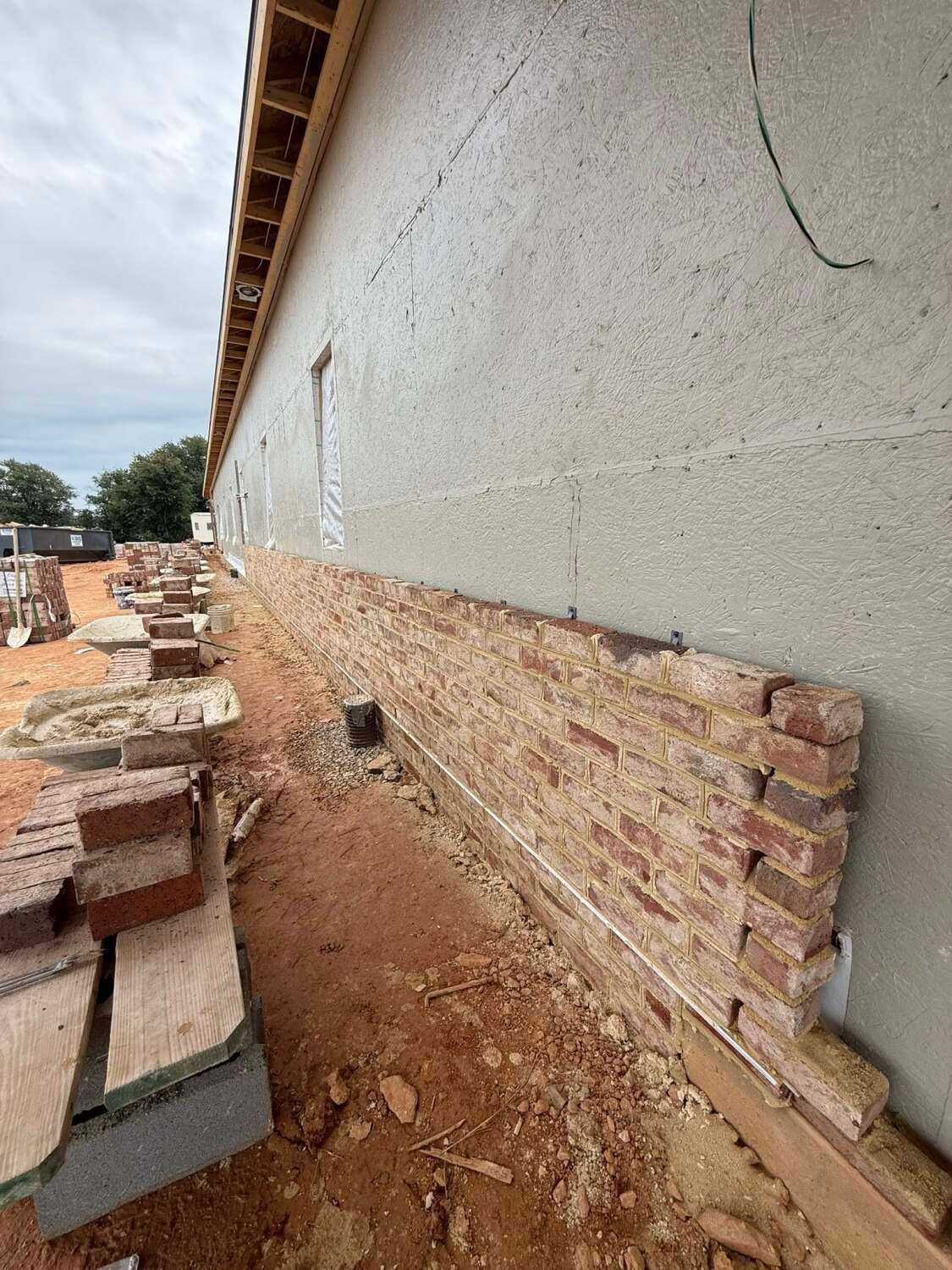 Building exterior under construction; brick veneer installation along the wall's base. Reddish-brown bricks, gray stucco.