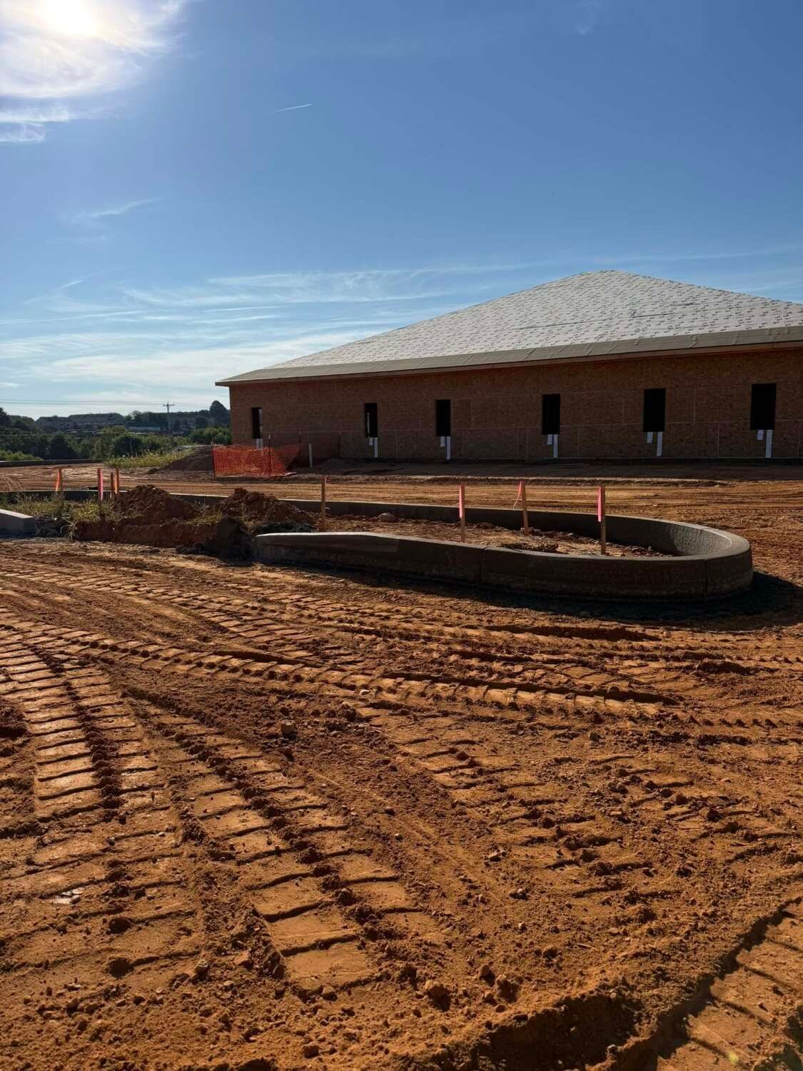 Construction site with a brick building under a blue sky, dirt, and tire tracks.