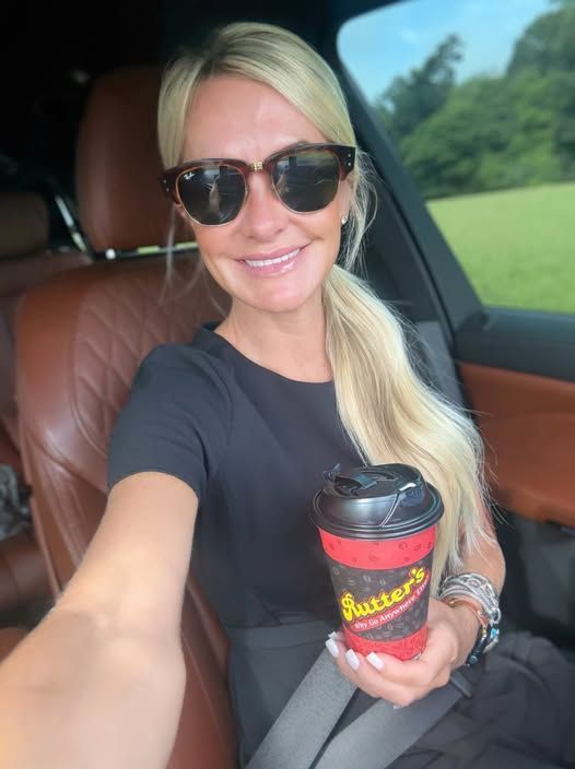 Woman in sunglasses, holding a coffee cup, smiling in a car. Brown leather seats.