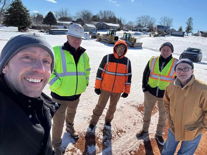 Group of five people posing for a selfie in a snowy construction site. Four are wearing safety vests and hats. A bulldozer is visible.