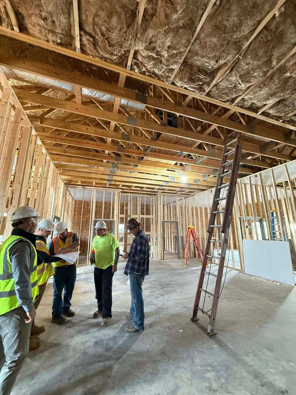 Construction workers in a building under construction, discussing plans. Wooden framework and ladder visible.