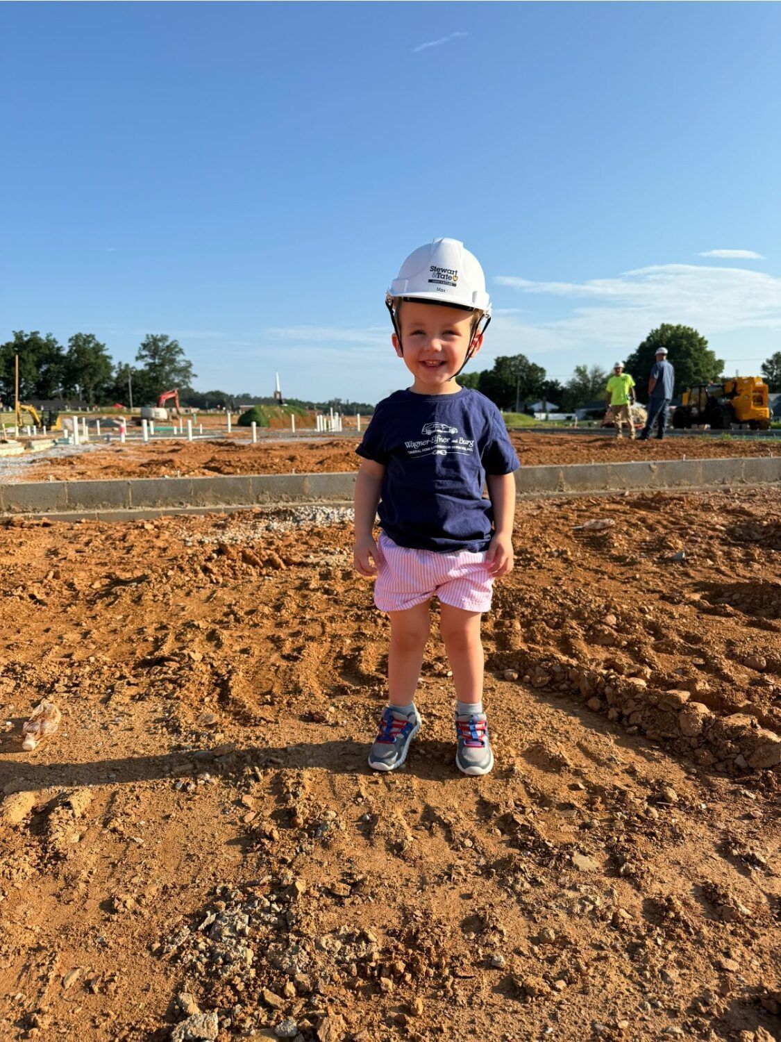 Child wearing a hard hat smiles on a construction site; brown dirt, blue sky.