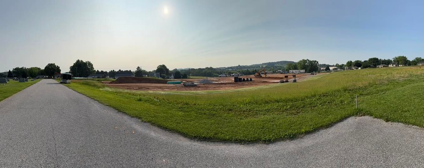 A gravel road leads past a grassy field and construction site under a bright sky.