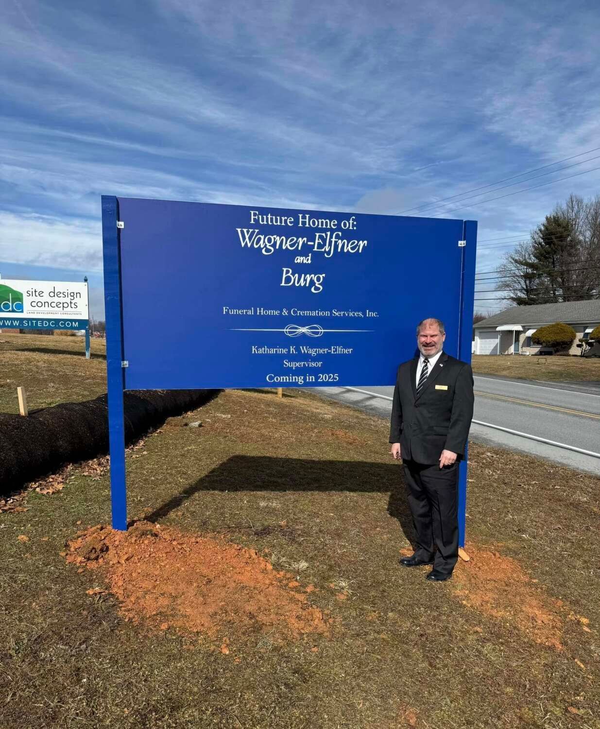 Man stands next to a blue sign for Wagner-Elfer and Baag. Sign on a sunny day.