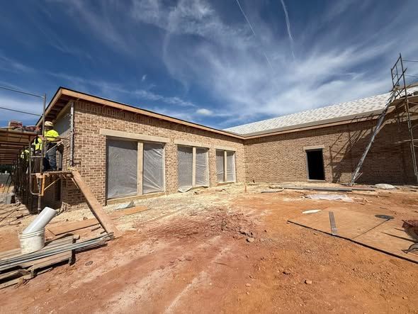 Brick building under construction, workers on scaffolding. Reddish-brown dirt, blue sky with wispy clouds.