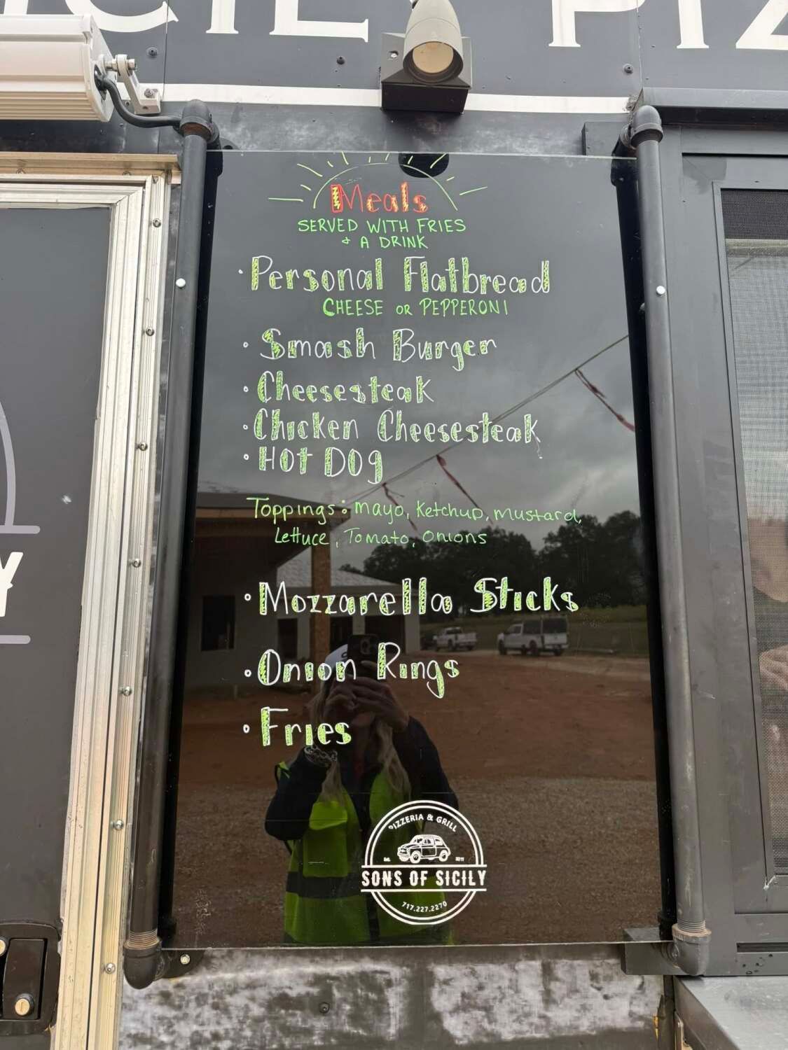 Menu board on a food truck with items like burgers, cheesesteak, and fries, with a reflection of a person wearing a safety vest.