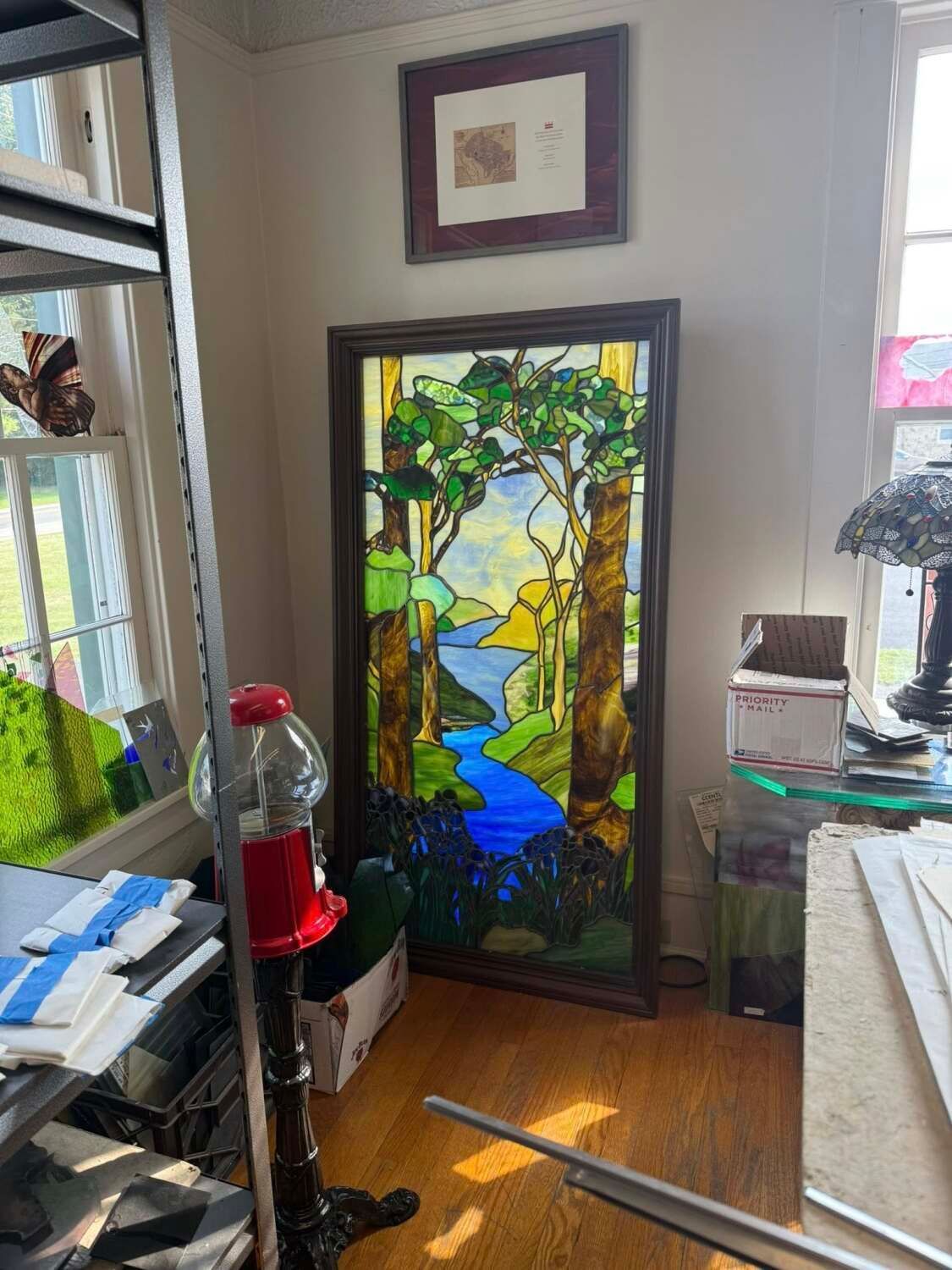 Stained glass window depicting a forest scene with a blue river, framed and leaning against a wall.