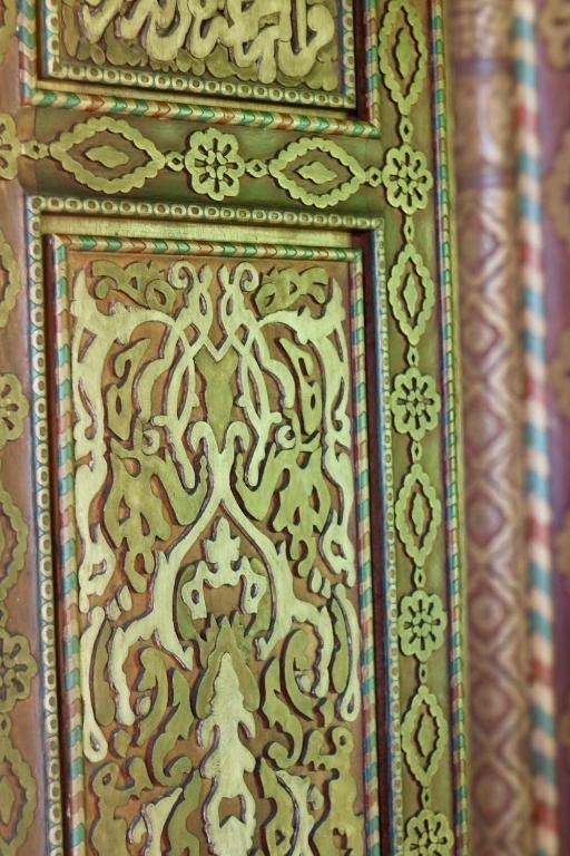 Primer plano de un panel de puerta tallado y pintado con intrincados diseños florales y geométricos en tonos verde y dorado.
