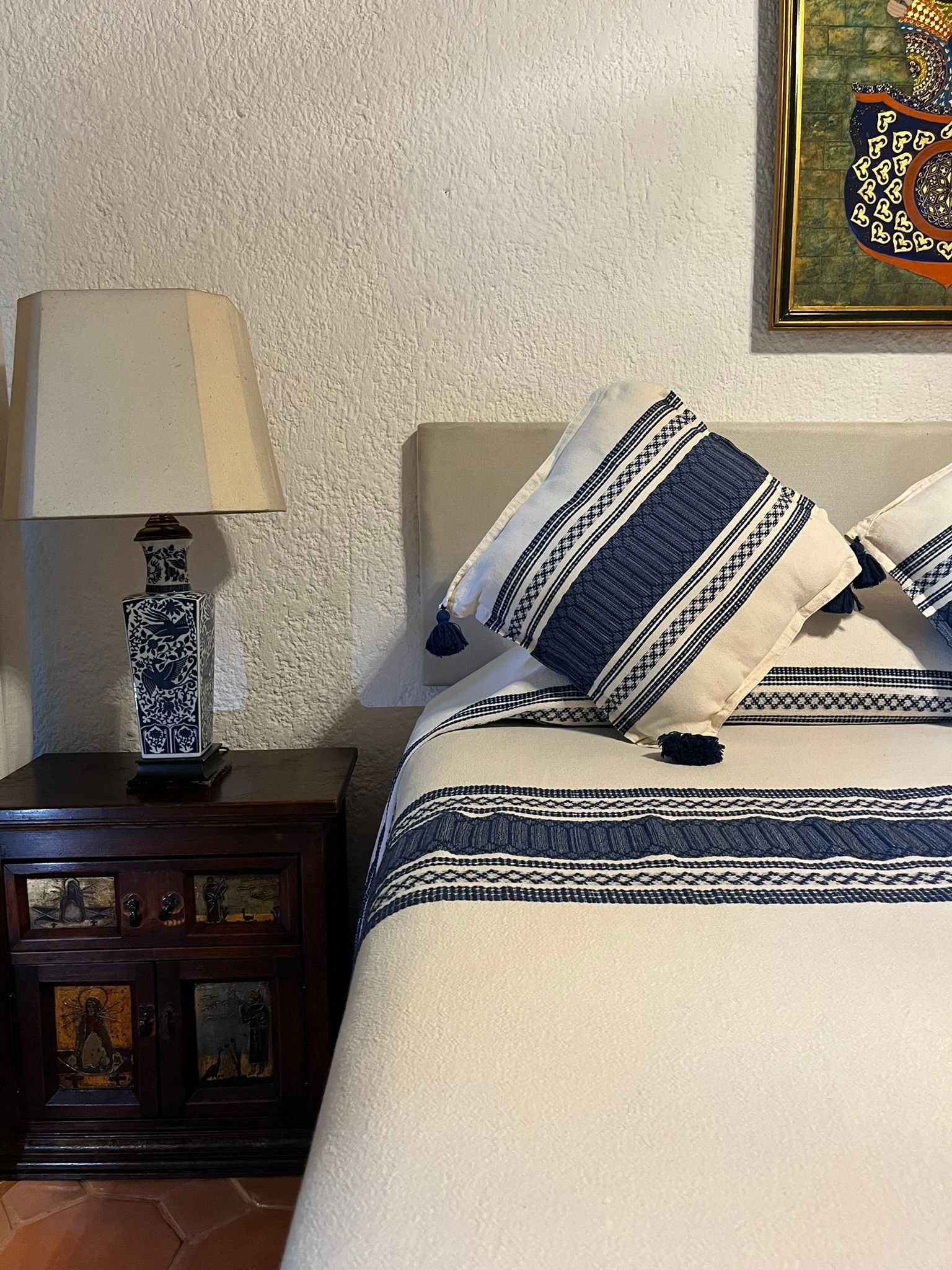 Dormitorio con colcha estampada en azul y blanco, almohadas, lámpara y mesita de noche de madera con un cuadro.