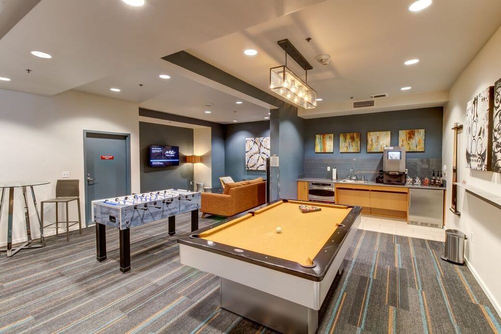 A recreational room with a pool table, a foosball table, a kitchenette, and lounge seating on a patterned gray carpet.