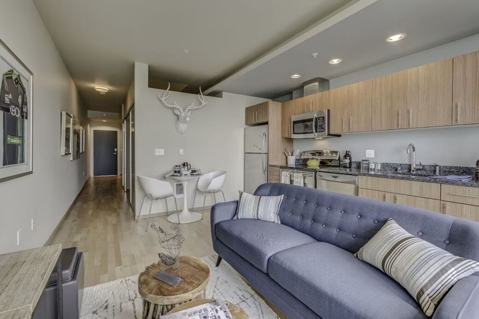 Modern studio apartment with a gray tufted sofa, wooden kitchen cabinets, a small dining table, and a hallway entrance.