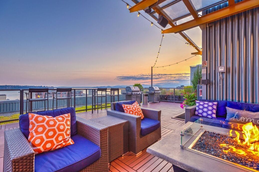 Rooftop community lounge with blue cushioned seating, a fire pit, string lights, and a water view.