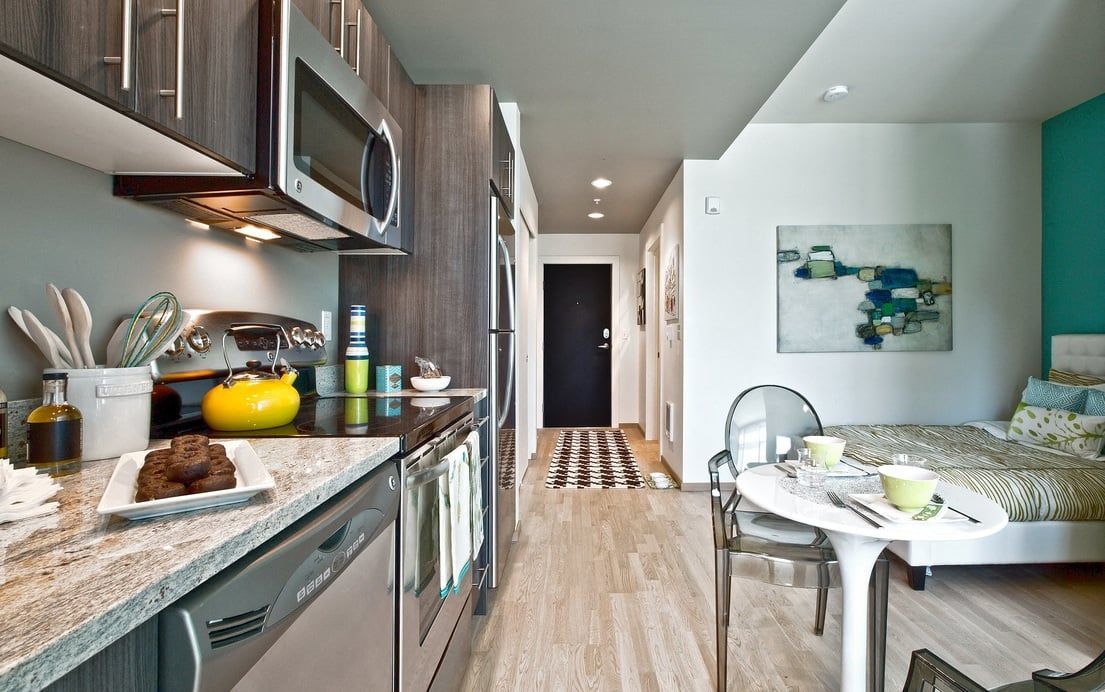 Open-plan kitchen with stainless-steel appliances; dining table and bed visible in living area.