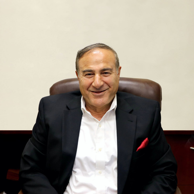 Man in a suit smiles, seated in a brown chair, red pocket square visible. Plain beige background.