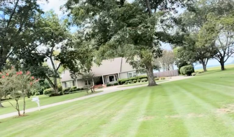 A house with a long driveway and well-manicured green lawn, framed by trees under a blue sky.