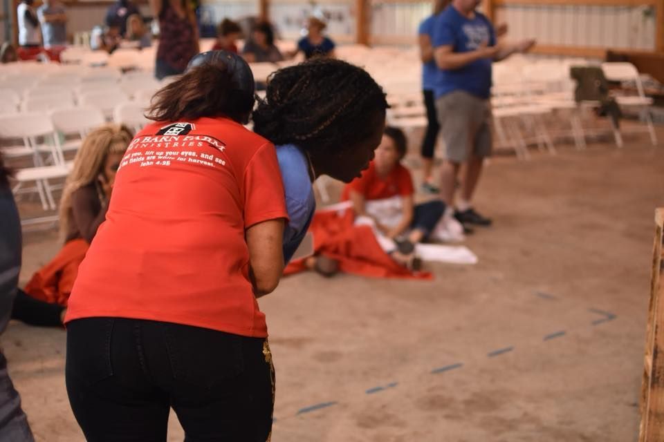 Two women in a large room, one in red shirt assisting the other. Others in background.
