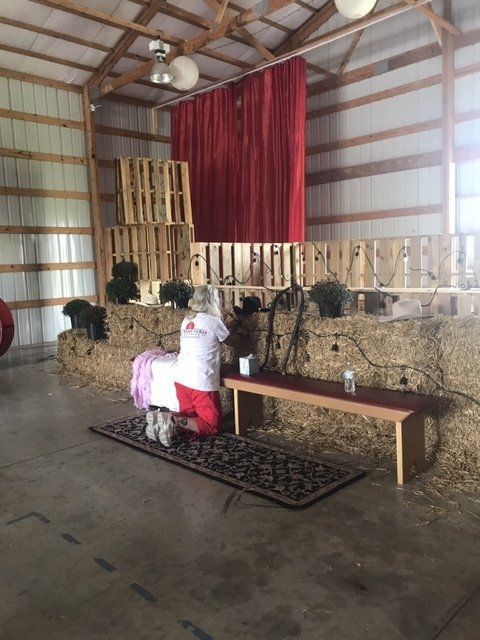 A woman kneels, praying, on a rug in a barn with hay bales, wooden pallets, and a red curtain.